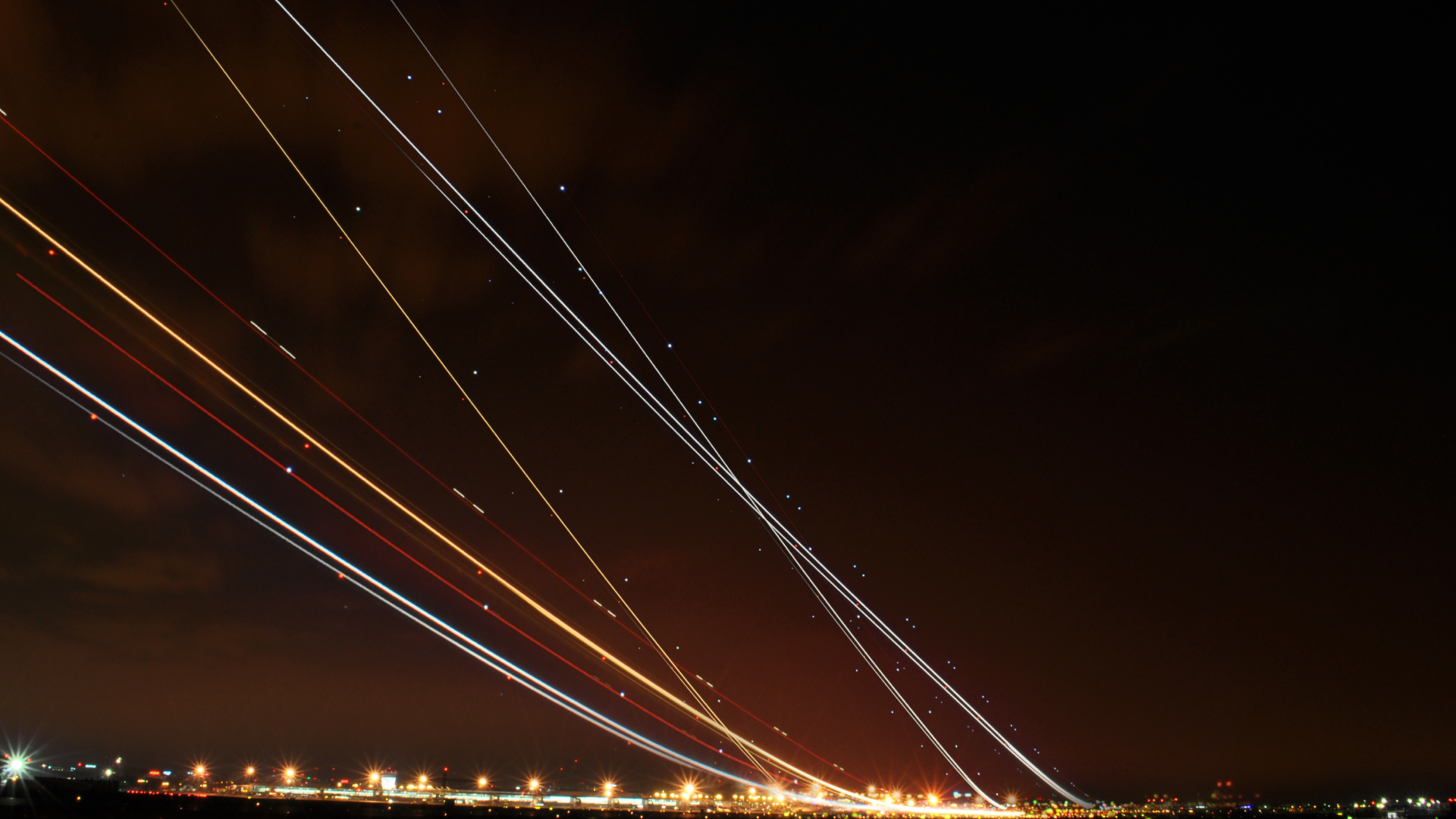 Long-exposure image of aircraft flight paths streaking across the night sky above a brightly lit airport, illustrating high-volume air traffic coordination.